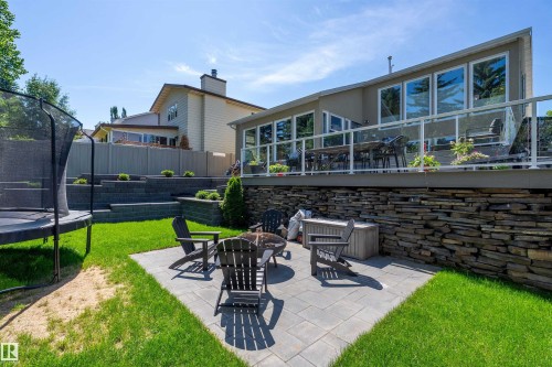 Expansive backyard featuring a paver patio with fire pit, terraced retaining walls, a raised composite deck with glass panel railing, and a stacked stone base - 4 Wentworth Crescent, St. Albert, AB 