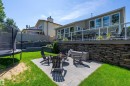 Expansive backyard featuring a paver patio with fire pit, terraced retaining walls, a raised composite deck with glass panel railing, and a stacked stone base - 4 Wentworth Crescent, St. Albert, AB 