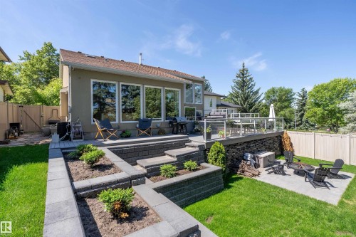 Multi-level backyard featuring a spacious upper deck with glass railings, a lower patio area with a built-in fire pit, tiered retaining walls with integrated planters, and a manicured lawn - 4 Wentworth Crescent, St. Albert, AB 