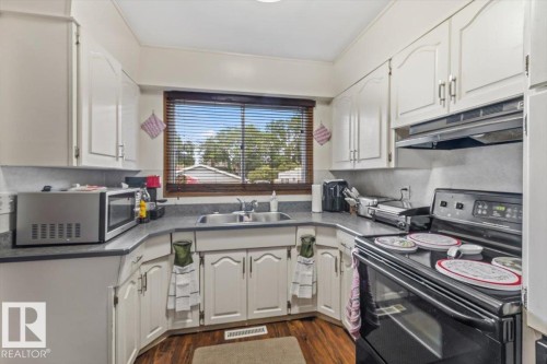 Functional kitchen featuring white cabinetry, dark countertops, a double basin sink beneath a window, and wood-finish flooring - 4622 Ada Boulevard, Edmonton, AB 