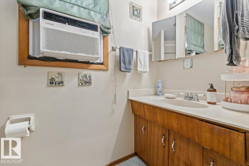 Bathroom featuring a window-mounted air conditioning unit, a wood-finish vanity with an integrated sink, a light-toned countertop, and an overhead mirror - 4622 Ada Boulevard, Edmonton, AB 