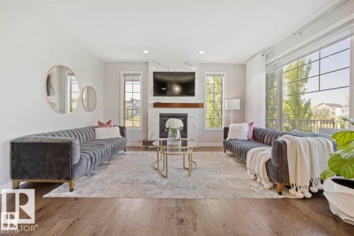 Spacious living area featuring large windows, a central fireplace with a white marble-look surround, and wood-finish flooring - 1004 Huckell Pl Sw, Edmonton, AB - Indoor Photo Showing Living Room With Fireplace