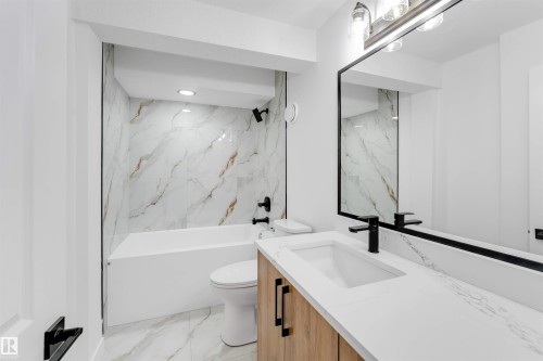 Bathroom featuring a white soaking tub, marble-finish wall and floor tile, a light wood-finish vanity with a white countertop, and a contemporary frameless mirror - 5512 150 Avenue, Edmonton, AB 