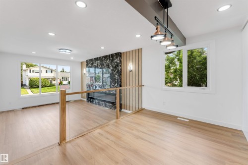 Open-concept living area featuring light wood-finish flooring, a modern fireplace with a dark marbled surround, and a glass-paneled railing - 5512 150 Avenue, Edmonton, AB 