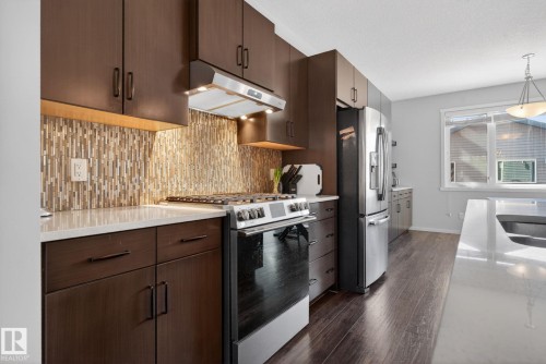 Contemporary kitchen featuring dark wood cabinetry, stainless steel appliances, a mosaic tile backsplash, light countertops, and wood-finish flooring - 3456 Cutler Crescent, Edmonton, AB - Indoor Photo Showing Kitchen With Upgraded Kitchen