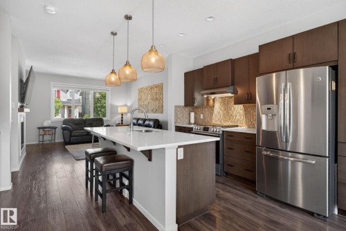 Contemporary kitchen featuring a white quartz island countertop with seating, stainless steel appliances, dark wood-finish cabinetry, and a textured backsplash, opening to a living area with wood-finish flooring - 3456 Cutler Crescent, Edmonton, AB - Indoor Photo Showing Kitchen With Upgraded Kitchen