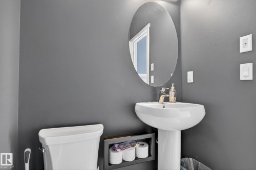 Pedestal sink with a gold-tone faucet and an oval mirror, complemented by a white toilet and dark gray walls - 3456 Cutler Crescent, Edmonton, AB - Indoor Photo Showing Bathroom