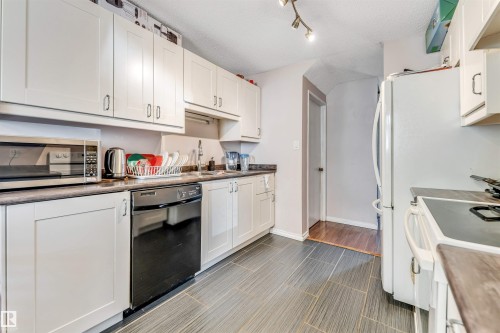 Kitchen featuring white cabinetry with brushed nickel hardware, dark countertops, a black dishwasher, and grey plank-style flooring - 24 1651 46 Street, Edmonton, AB 