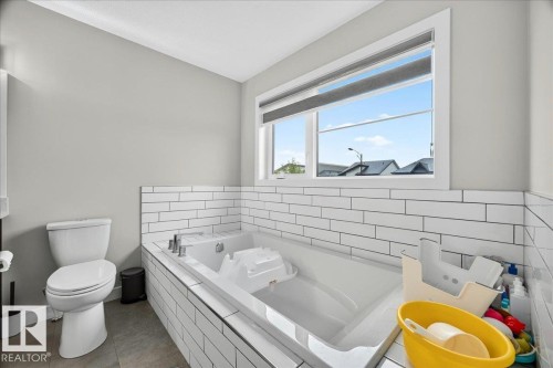 Soaking tub with white subway tile surround, white toilet, light gray walls, window with roller shade, and gray tile flooring - 18139 75 Street, Edmonton, AB 