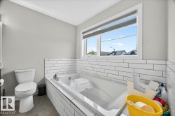 Soaking tub with white subway tile surround, white toilet, light gray walls, window with roller shade, and gray tile flooring - 