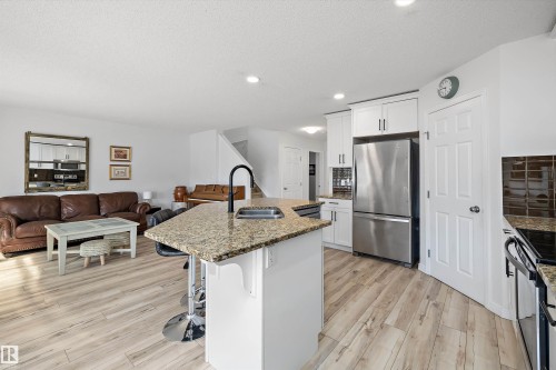 Open-concept living space featuring wood-finish flooring, a kitchen island with stone countertop, white shaker cabinetry, and stainless steel appliances - 25 Halladay Boulevard, Spruce Grove, AB - Indoor Photo Showing Kitchen With Double Sink With Upgraded Kitchen