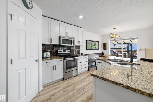 Kitchen featuring a granite countertop island with an undermount sink, stainless steel appliances, white cabinetry with dark hardware, wood-finish flooring, and a sliding glass door leading to an outdoor space - 25 Halladay Boulevard, Spruce Grove, AB - Indoor Photo Showing Kitchen With Double Sink With Upgraded Kitchen