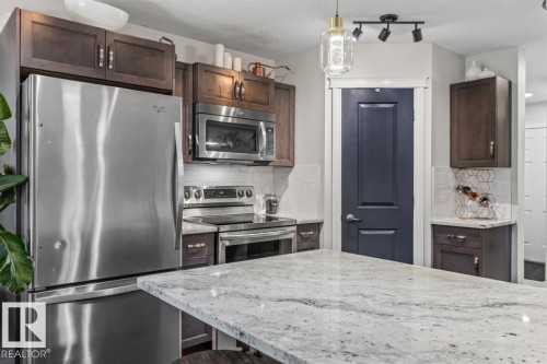 Kitchen featuring a granite-look island, stainless steel appliances, dark wood cabinetry, a tile backsplash, and a paneled door - 14 Edinburgh Crt N, St. Albert, AB 