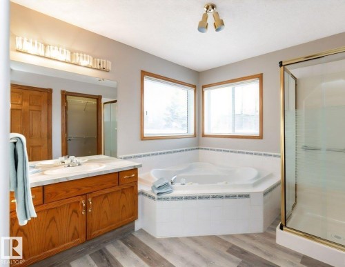 Bathroom featuring a corner jetted tub, dual windows with wood trim, a glass-enclosed shower, a wood-finish vanity with a single sink and light-colored countertop, and wood-finish flooring - 49 Heritage Drive, St. Albert, AB 