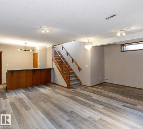 Spacious interior featuring wood-finish flooring, a built-in wet bar with a dark countertop, a staircase with wood railing and carpeted treads, and recessed lighting - 49 Heritage Drive, St. Albert, AB 