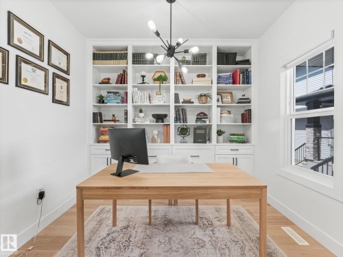 Dedicated office space featuring extensive built-in shelving and cabinetry, natural wood-finish flooring, a modern chandelier with exposed bulbs, and a large window providing natural light - 5411 66 Street, Beaumont, AB 