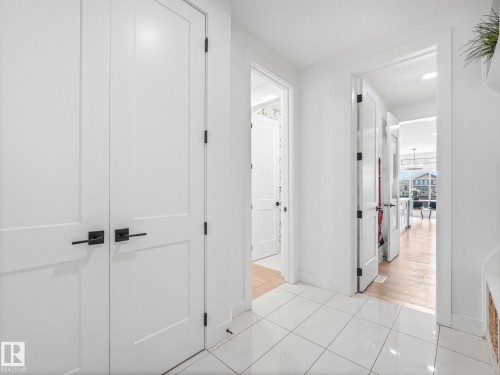 Hallway featuring white panel doors with matte black hardware, white square tile flooring, and light wood-finish flooring visible through doorways - 5411 66 Street, Beaumont, AB 