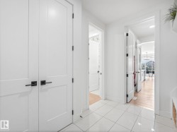 Hallway featuring white panel doors with matte black hardware, white square tile flooring, and light wood-finish flooring visible through doorways - 