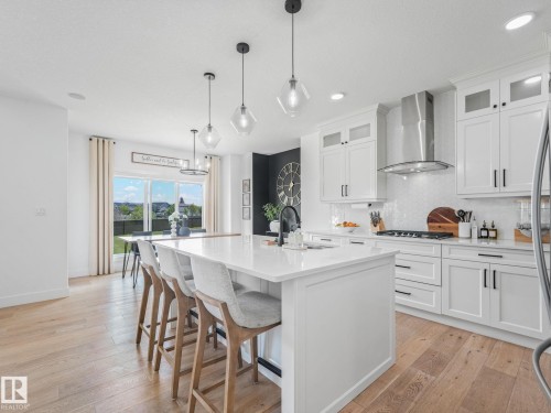 White kitchen featuring an expansive center island with seating, a gas range with stainless steel hood, shaker-style cabinetry, and wood-finish flooring - 5411 66 Street, Beaumont, AB 