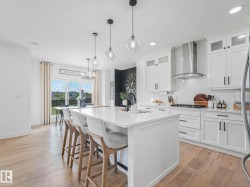 White kitchen featuring an expansive center island with seating, a gas range with stainless steel hood, shaker-style cabinetry, and wood-finish flooring - 