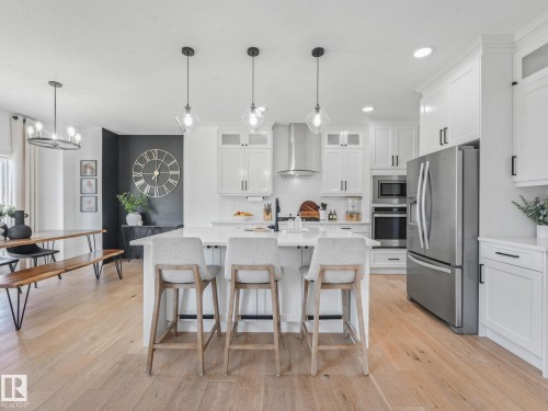 Open-concept kitchen featuring white cabinetry, stainless steel appliances, a central island with seating, pendant lighting, and wood-finish flooring - 5411 66 Street, Beaumont, AB 