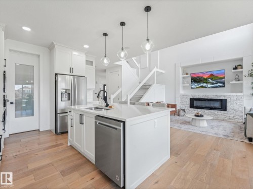 Open-concept living area featuring a kitchen island with pendant lighting, stainless steel appliances, wood-finish flooring, a white stone fireplace surround, and built-in shelving - 5411 66 Street, Beaumont, AB 