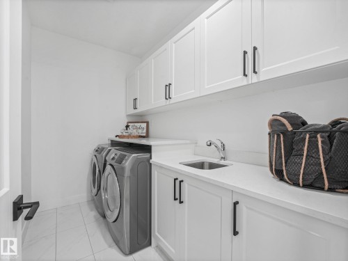 Laundry area featuring white shaker-style cabinetry with matte black hardware, a stainless steel sink with a chrome faucet, and light-toned solid surface countertops - 5411 66 Street, Beaumont, AB 