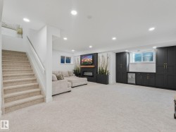 Spacious lower-level living area featuring recessed lighting, plush carpet flooring, a built-in media center with an electric fireplace, and extensive dark-toned cabinetry with a beverage refrigerator - 