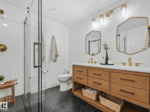 Contemporary bathroom featuring a glass-enclosed shower with white geometric tile, a dual vanity with a light wood-finish cabinet, white countertops, and black hexagonal floor tiles - 5411 66 Street, Beaumont, AB 