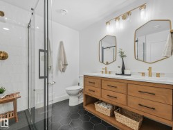 Contemporary bathroom featuring a glass-enclosed shower with white geometric tile, a dual vanity with a light wood-finish cabinet, white countertops, and black hexagonal floor tiles - 