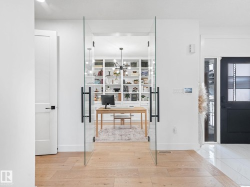 Glass French doors with matte black hardware open to a room featuring extensive built-in shelving, a contemporary chandelier, and wood-finish flooring - 5411 66 Street, Beaumont, AB 