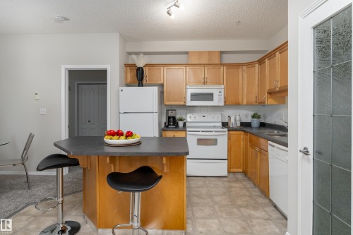 Kitchen featuring light wood cabinetry, white appliances, a dark countertop, and an angled peninsula with seating - 404 12111 51 Avenue, Edmonton, AB 
