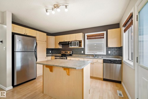 Kitchen featuring light wood-finish cabinetry, stainless steel appliances, a central island with countertop, dark subway tile backsplash, and wood-finish flooring - 2369 27 Avenue, Edmonton, AB 