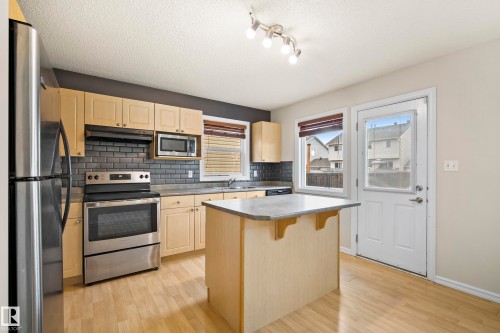 Contemporary kitchen featuring stainless steel appliances, light wood-finish cabinetry, a dark subway tile backsplash, and a central island with a contrasting countertop - 2369 27 Avenue, Edmonton, AB 