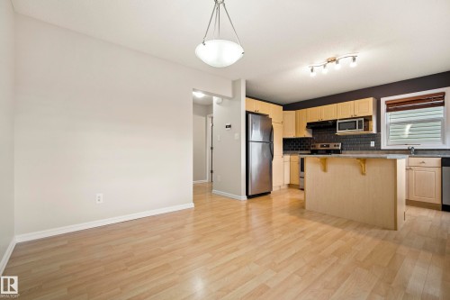 Open-concept kitchen and dining area featuring light wood-finish flooring, a kitchen island with seating, stainless steel appliances, and a dark tile backsplash - 2369 27 Avenue, Edmonton, AB 