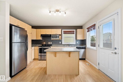Kitchen featuring wood-finish flooring, light wood cabinetry, stainless steel appliances, a center island with a light-toned countertop, and a white exterior door with a glass insert - 2369 27 Avenue, Edmonton, AB 