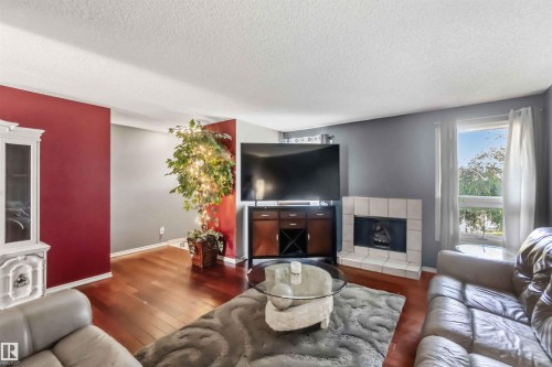Living room featuring wood-finish flooring, a white brick-surround fireplace, and a large window with natural lighting - 14121 23 Street, Edmonton, AB 