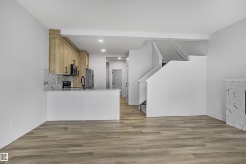 Open-concept living space featuring wood-finish flooring, a contemporary kitchen with light wood cabinetry and a white countertop, and a modern fireplace with a patterned surround - 1915 164 Street, Edmonton, AB 