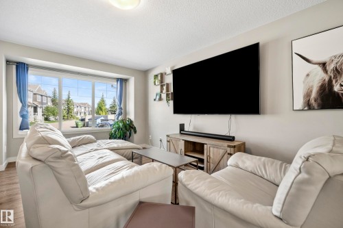 Spacious living area featuring a large bay window, light wood-finish flooring, a contemporary media console with barn door accents, and recessed ceiling lighting - 11 4205 30 Street, Edmonton, AB 