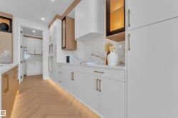 Contemporary kitchen featuring herringbone wood-finish flooring, white flat-panel cabinetry, a full-height backsplash, gold-finish hardware, and integrated under-cabinet lighting - 