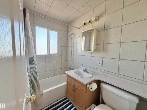 Bathroom featuring a built-in tub with shower, white wall tiling, a vanity with wood-finish cabinetry, a built-in sink, and a mirrored medicine cabinet - 11941 78 Street, Edmonton, AB 