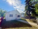 White siding residence featuring a front entry door with an arched glass insert, contrasting red trim around windows and door, a gravel pathway, a red wooden fence, and mature trees - 11941 78 Street, Edmonton, AB 