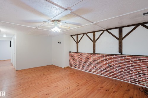 Spacious interior room featuring wood-finish flooring, a ceiling fan with integrated lighting, a brick-style accent wall, and decorative dark wood ceiling beams - 212 Homestead Crescent, Edmonton, AB 