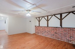 Spacious interior room featuring wood-finish flooring, a ceiling fan with integrated lighting, a brick-style accent wall, and decorative dark wood ceiling beams - 