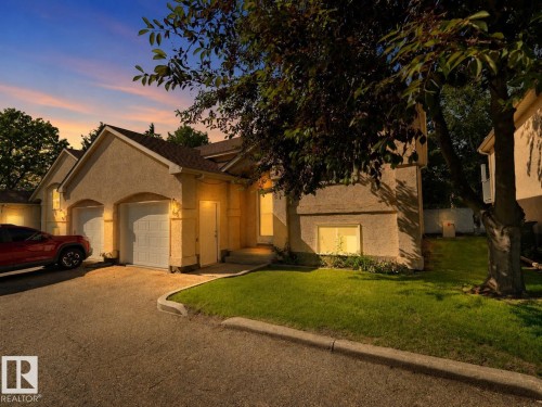 Stucco exterior residence featuring a two-car garage with arched entrances, a paved driveway, a landscaped lawn, and mature trees - 1855 Mill Woods Rd E Nw, Edmonton, AB 