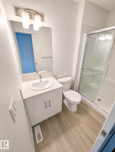 Contemporary bathroom featuring a white vanity with integrated sink, a glass-enclosed shower, a toilet, wood-finish flooring, and a modern vanity light fixture - 22845 84 Avenue, Edmonton, AB 