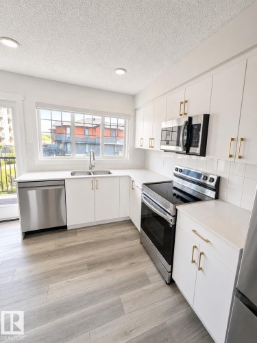 Modern kitchen featuring light wood-finish flooring, white cabinetry with gold-tone hardware, stainless steel appliances, and a white tile backsplash - 22845 84 Avenue, Edmonton, AB 