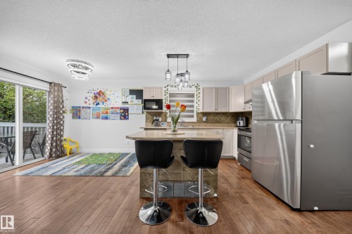 Open-concept kitchen and living area featuring wood-finish flooring, stainless steel appliances, a center island with seating, a tile backsplash, and a sliding glass door opening to an outdoor deck - 4 4350 23 Street, Edmonton, AB 