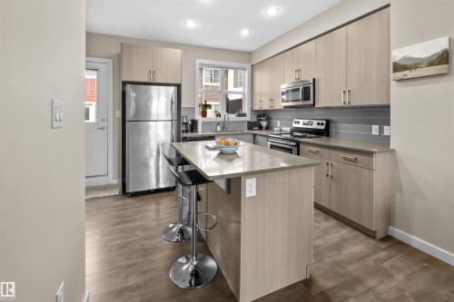 Contemporary kitchen featuring a central island with countertop seating, stainless steel appliances, light wood-finish cabinetry, and a grey subway tile backsplash - 13 2803 James Mowatt Trail, Edmonton, AB 