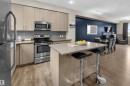 Open-concept kitchen featuring light wood-finish cabinetry, stainless steel appliances, a stone-finish island with seating, and wood-finish flooring - 13 2803 James Mowatt Trail, Edmonton, AB 